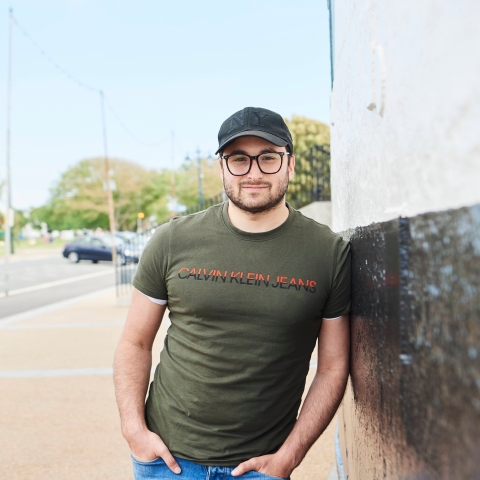 Male student in green top and cap with glasses leaning against wall