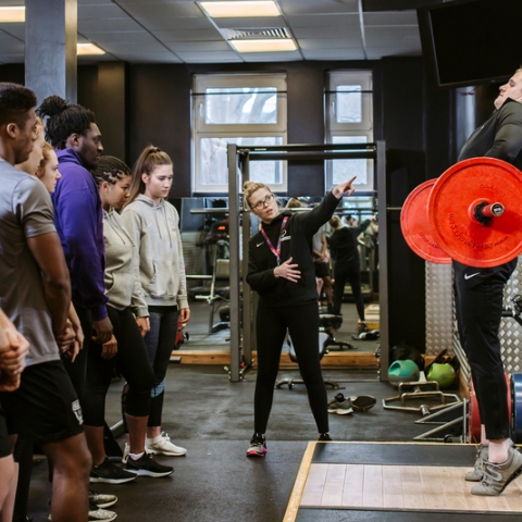  A group of students watching someone demonstrate lifting weights