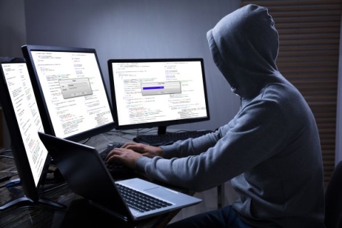 Person in dark room wearing a hoodie, sat in front of multiple computer screens